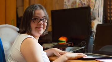 Young adult female with Down syndrome sitting in the bedroom of her home, using a laptop computer and smiling at the camera.