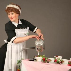 A lady in waitress outfit with a tea trolly pouring tea from a metal teapot.