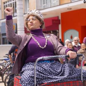 A man dressed as a grannie riding on a shopping trolley waving at the crowds.