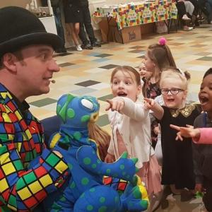 An entertainer in a bowler hat and brightly coloured jacket with a puppet frog entertaining a group of children.